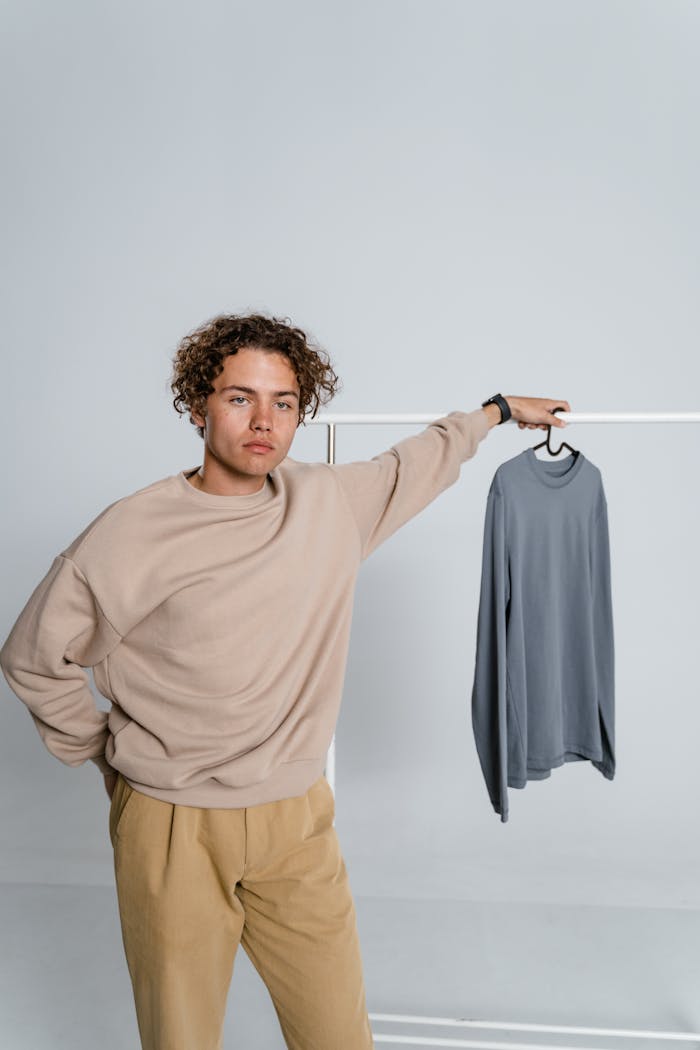 programs-02 Young man with curly hair posing in a minimalist studio with a sweater on a hanger.